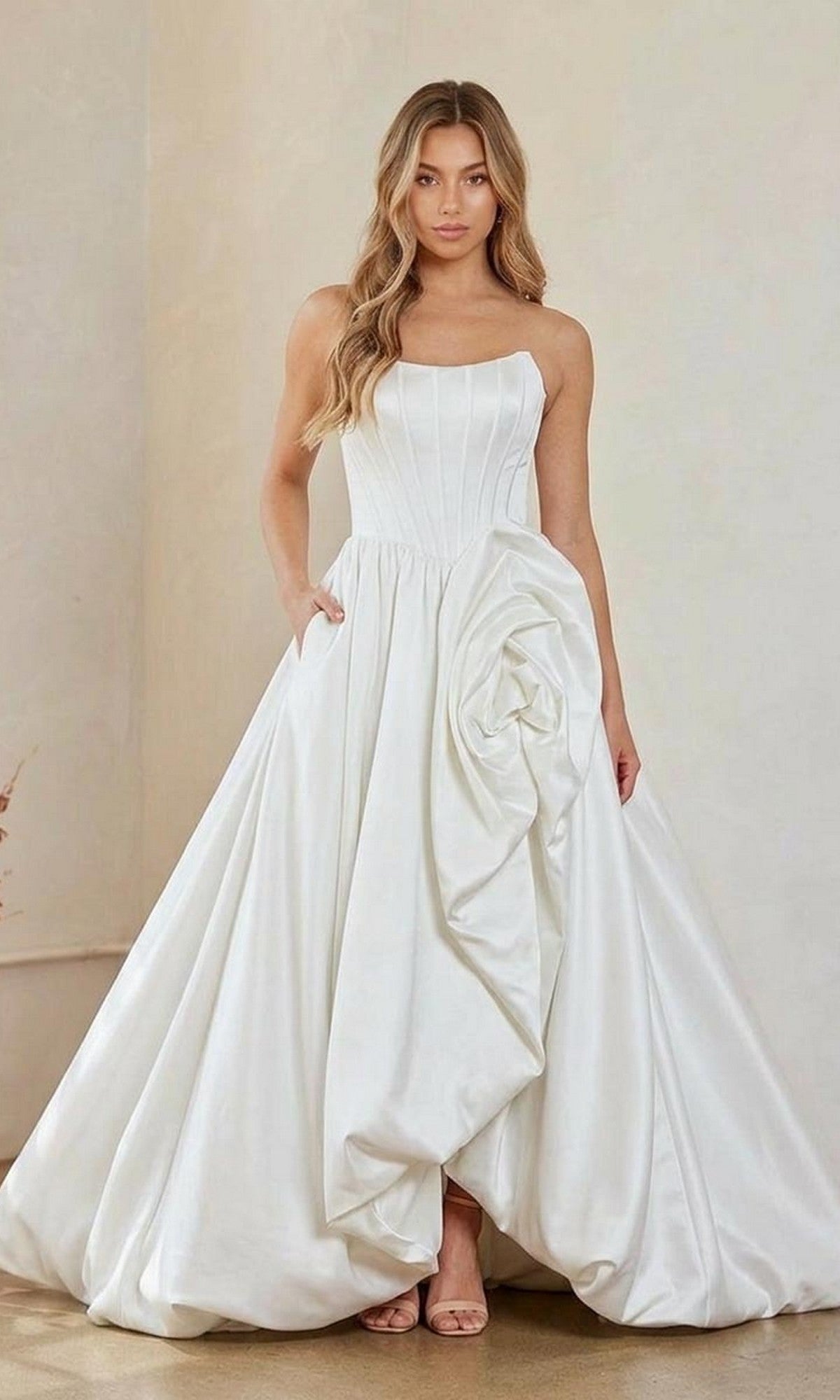 A woman wears the Amelia Couture 2085 long prom dress in white, a strapless gown with a structured corset bodice and a dramatic bubble-hem skirt featuring a large floral-inspired draped detail.